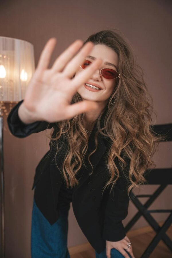 Attractive young female in stylish outfit and sunglasses looking at camera while standing with outstretched arm in modern studio with floor lamp