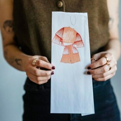 Crop faceless female dressmaker with tattoos on arms in trendy outfit demonstrating paper with fashion draft against white wall in studio