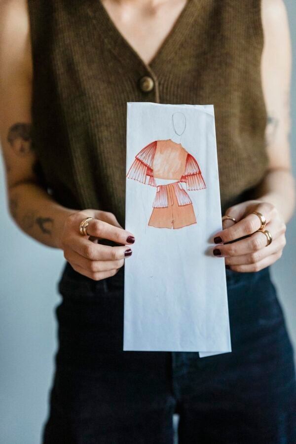 Crop faceless female dressmaker with tattoos on arms in trendy outfit demonstrating paper with fashion draft against white wall in studio