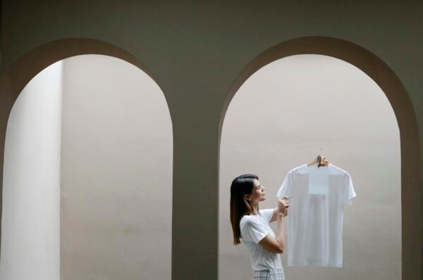 Side view of confident young lady with long dark hair in stylish outfit holding hanger with white t shire while standing near arch in minimalist house